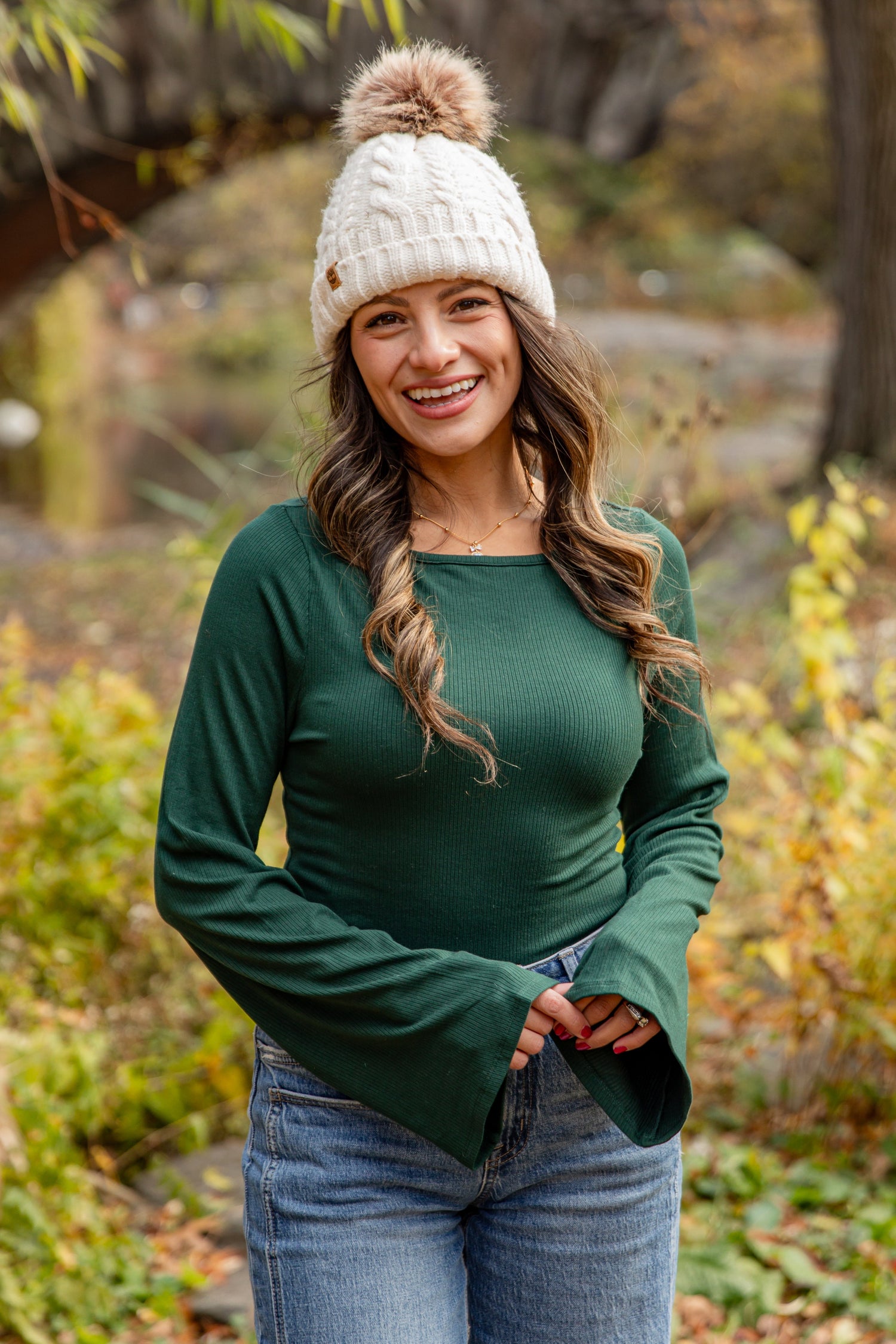 Woman wearing a green top and white knit hat in an autumn setting with trees and fallen leaves.
