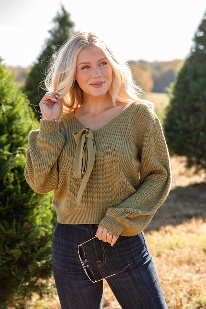 Woman wearing a mustard yellow sweater with a bow detail in a natural setting.
