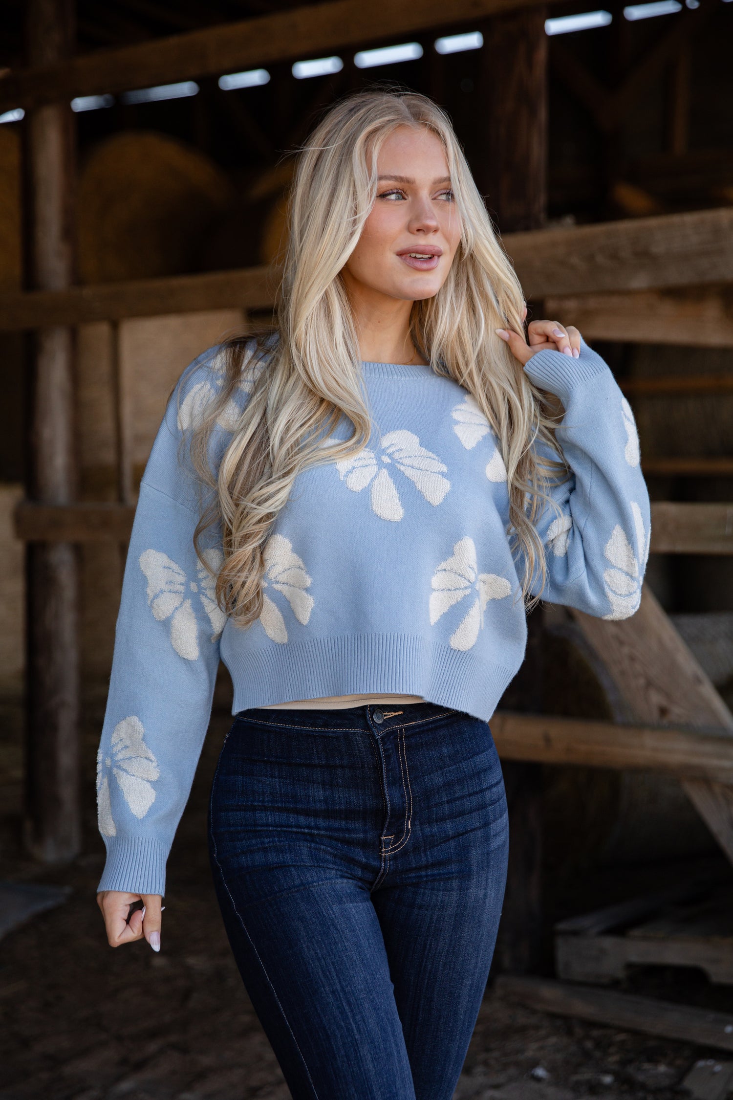 Woman wearing a light blue sweater with white floral patterns in a rustic setting.