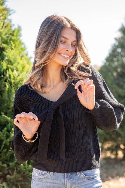 Woman wearing a black sweater and light blue jeans standing outdoors with trees in the background