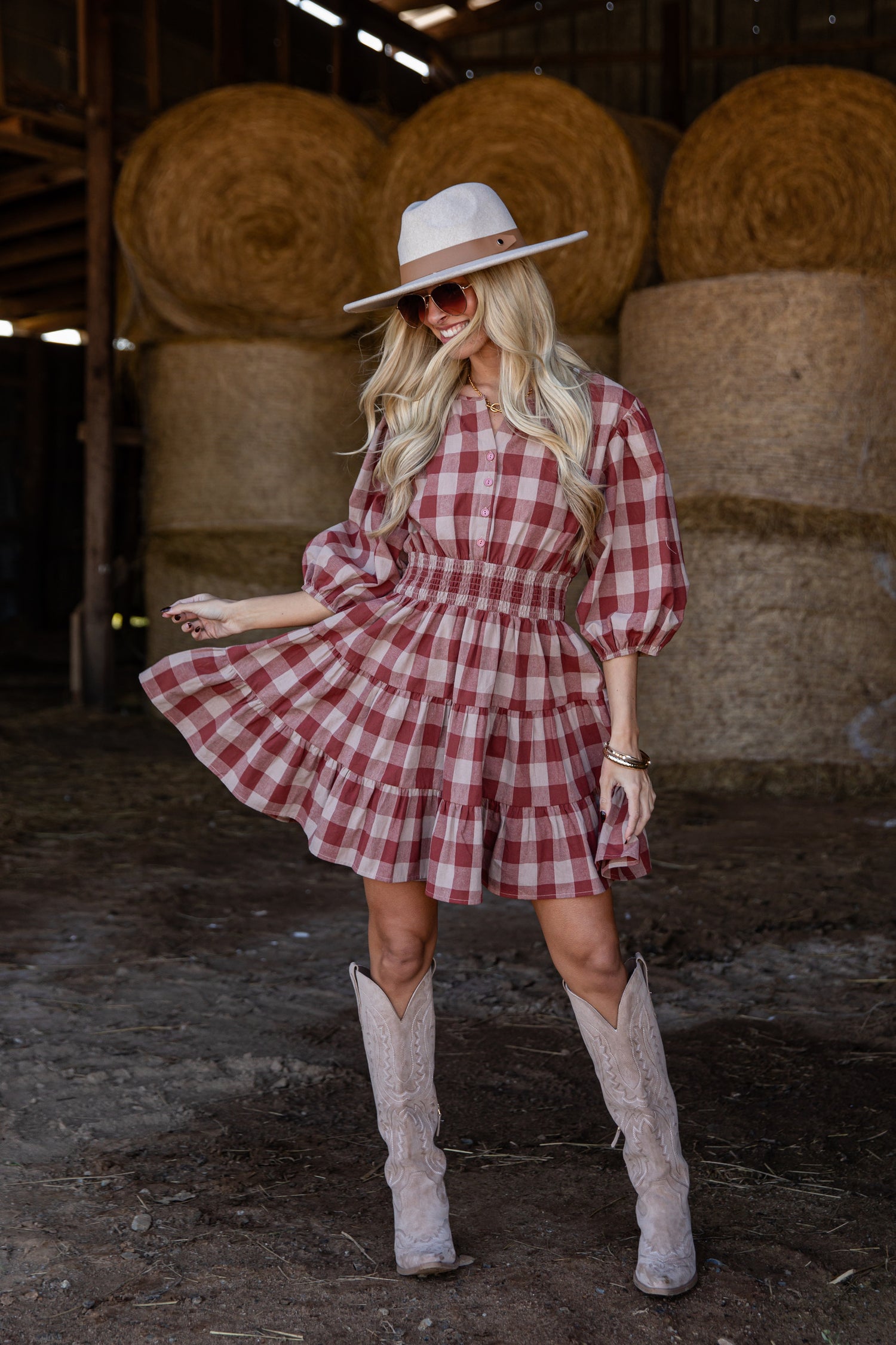 Woman in a red plaid dress and cowboy boots standing in a barn with hay bales.