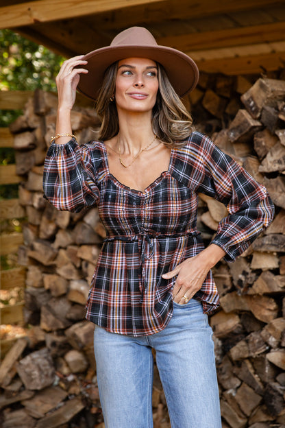 Woman wearing a plaid shirt and wide-brimmed hat standing in front of stacked firewood.