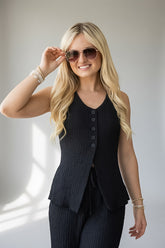 A blonde woman wearing a black ribbed sleeveless vest with front buttons and matching black ribbed pants, styled with sunglasses and bracelets, against a light gray background.