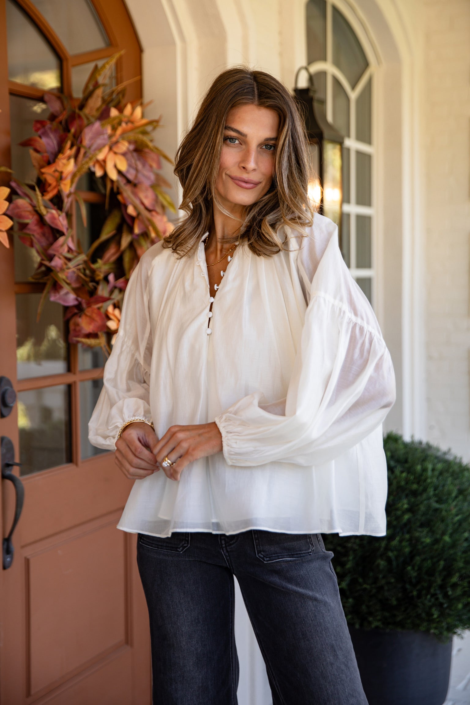 Woman wearing a white blouse and dark jeans standing in front of a door with floral decorations.