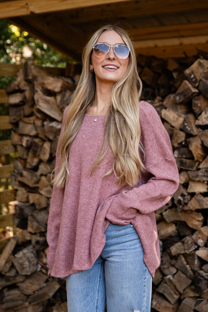 Woman wearing sunglasses and a pink sweater standing in front of stacked firewood.