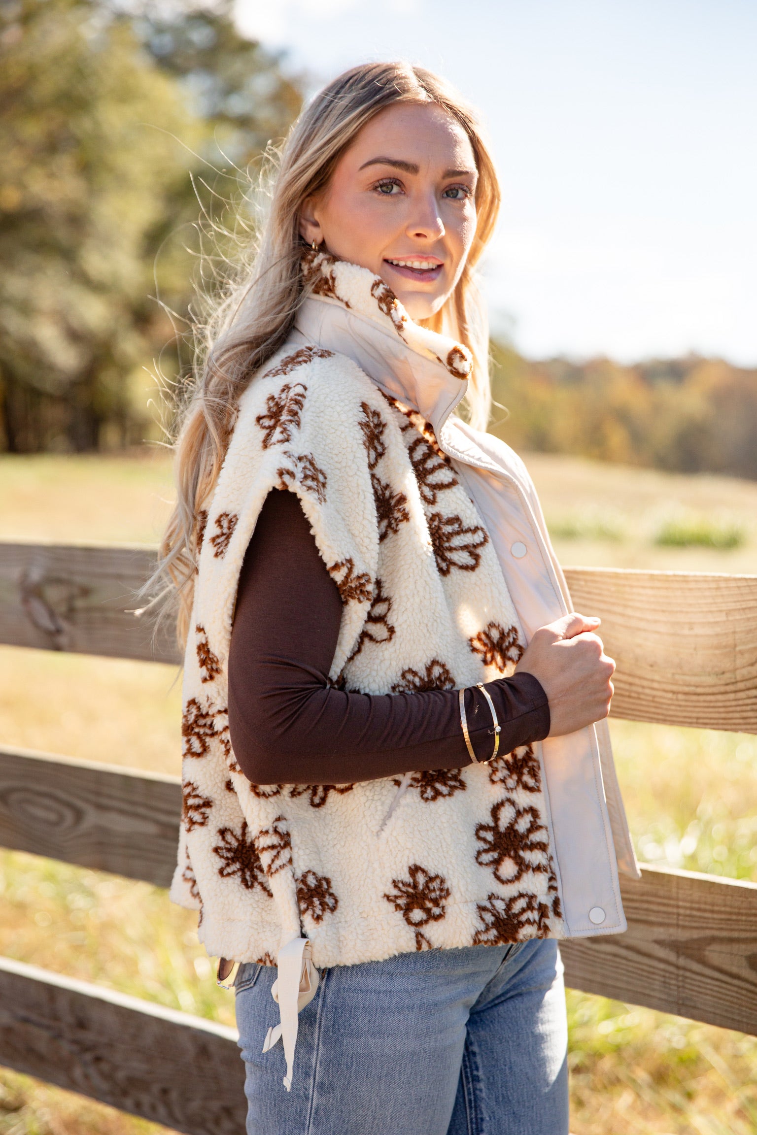 Woman wearing a white floral-patterned vest outdoors with a wooden fence and trees in the background.