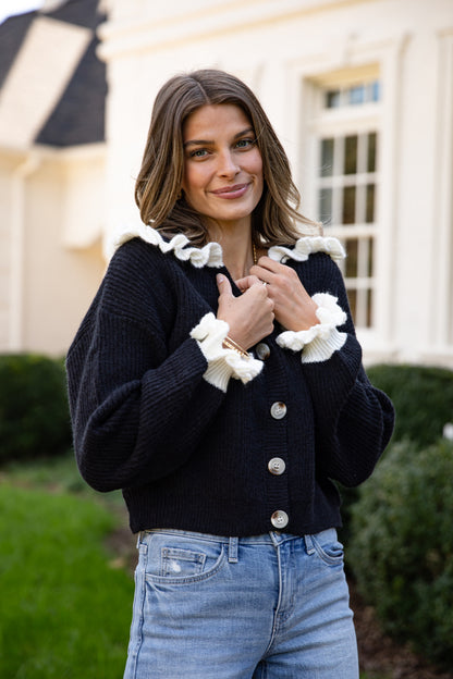 Woman wearing a black cardigan with white ruffled trim in front of a building.