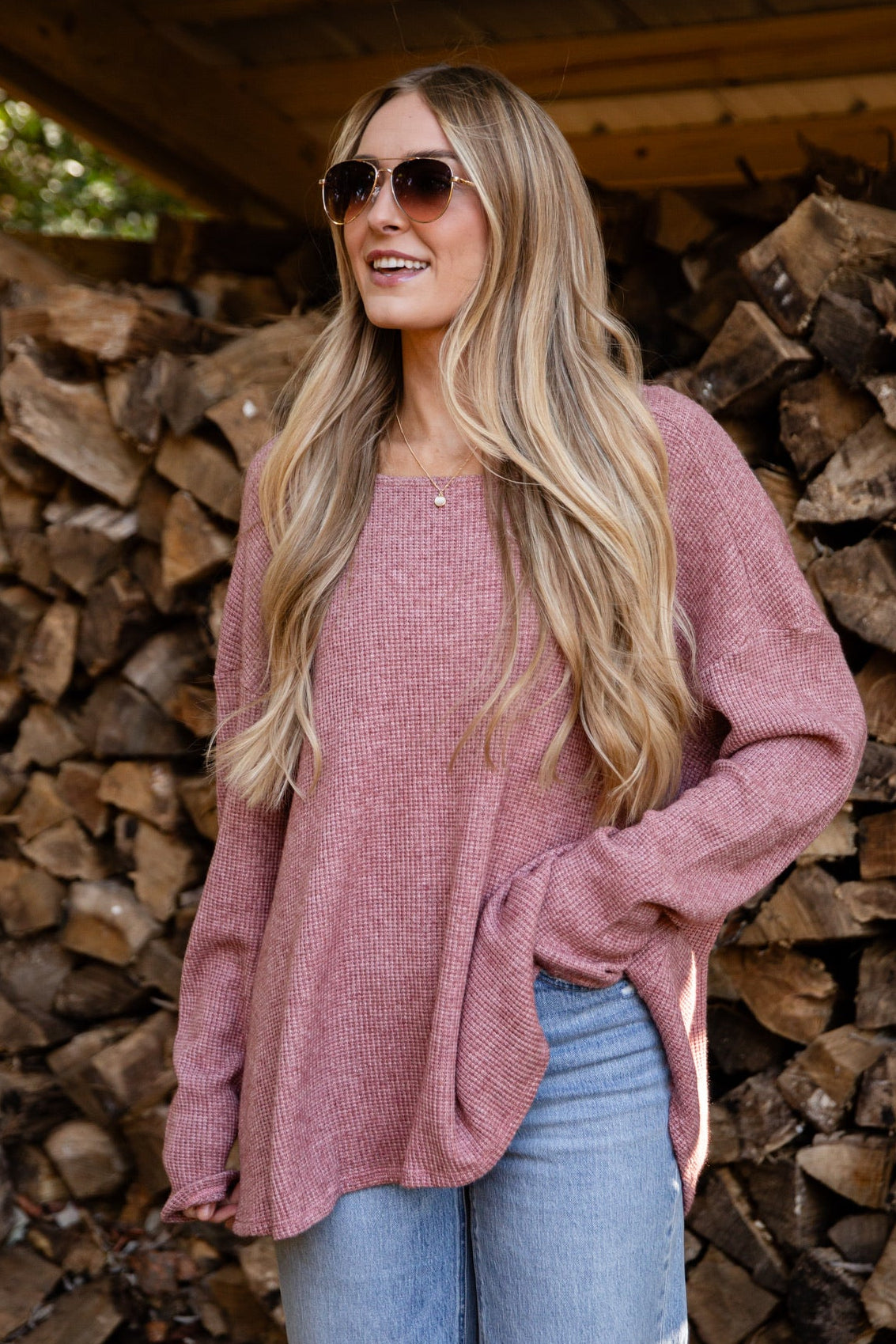Woman in a pink sweater and blue jeans standing in front of stacked firewood.