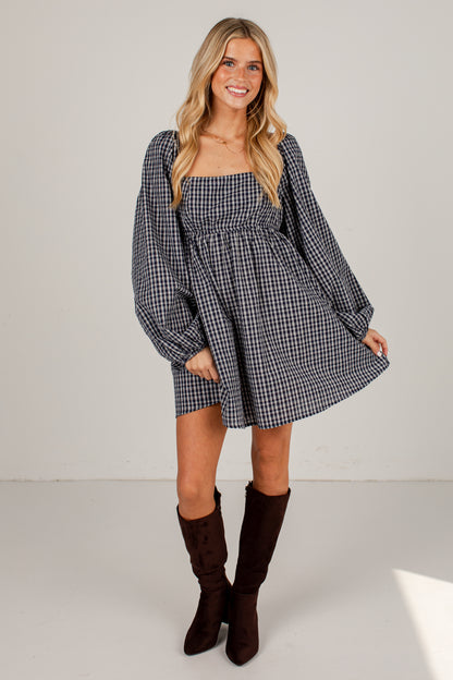 A woman modeling a black and white gingham mini dress with long balloon sleeves and a smocked bodice, paired with dark brown knee-high boots, against a plain light gray studio background.