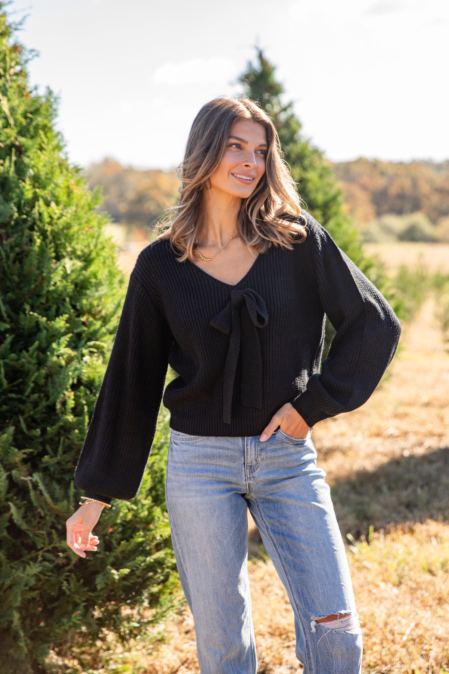 Woman wearing a black sweater and blue jeans standing in a field with trees in the background