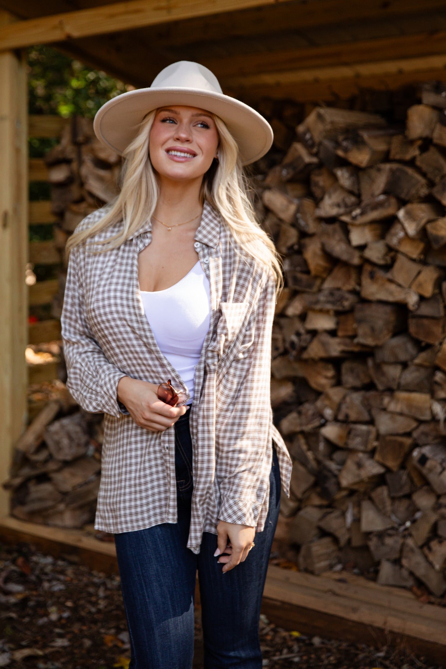 Woman wearing a plaid shirt, white top, and wide-brimmed hat standing in front of stacked firewood.