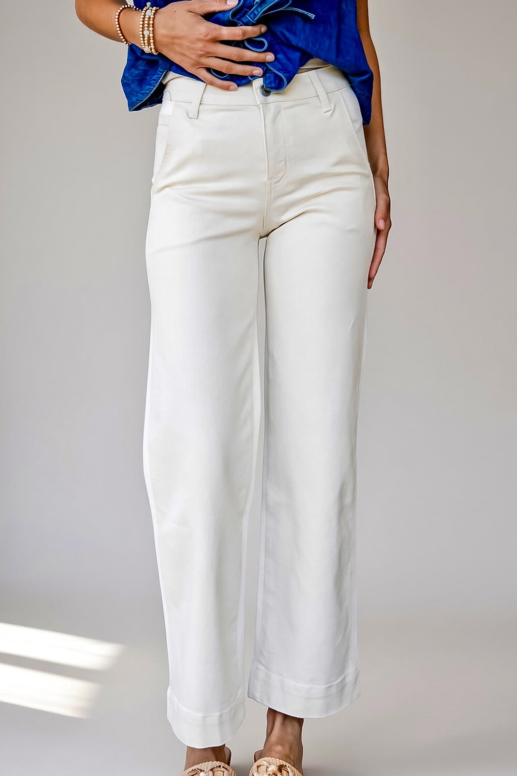 A person wearing white wide-leg cropped pants with a blue top and beige sandals, standing against a light gray studio background.