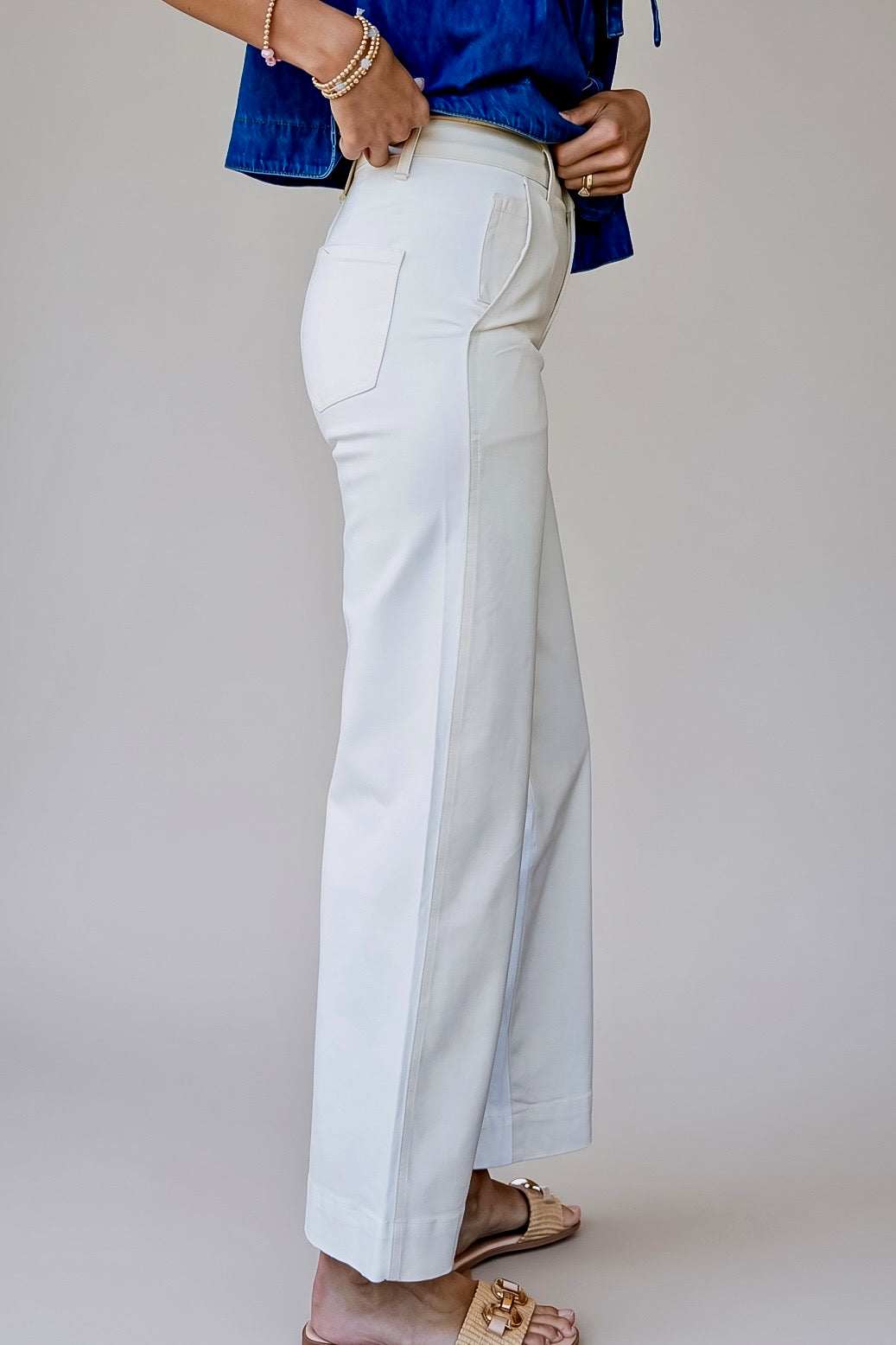 A person wearing white wide-leg cropped pants with a blue top and beige sandals, shown from the waist down against a plain light gray studio background.
