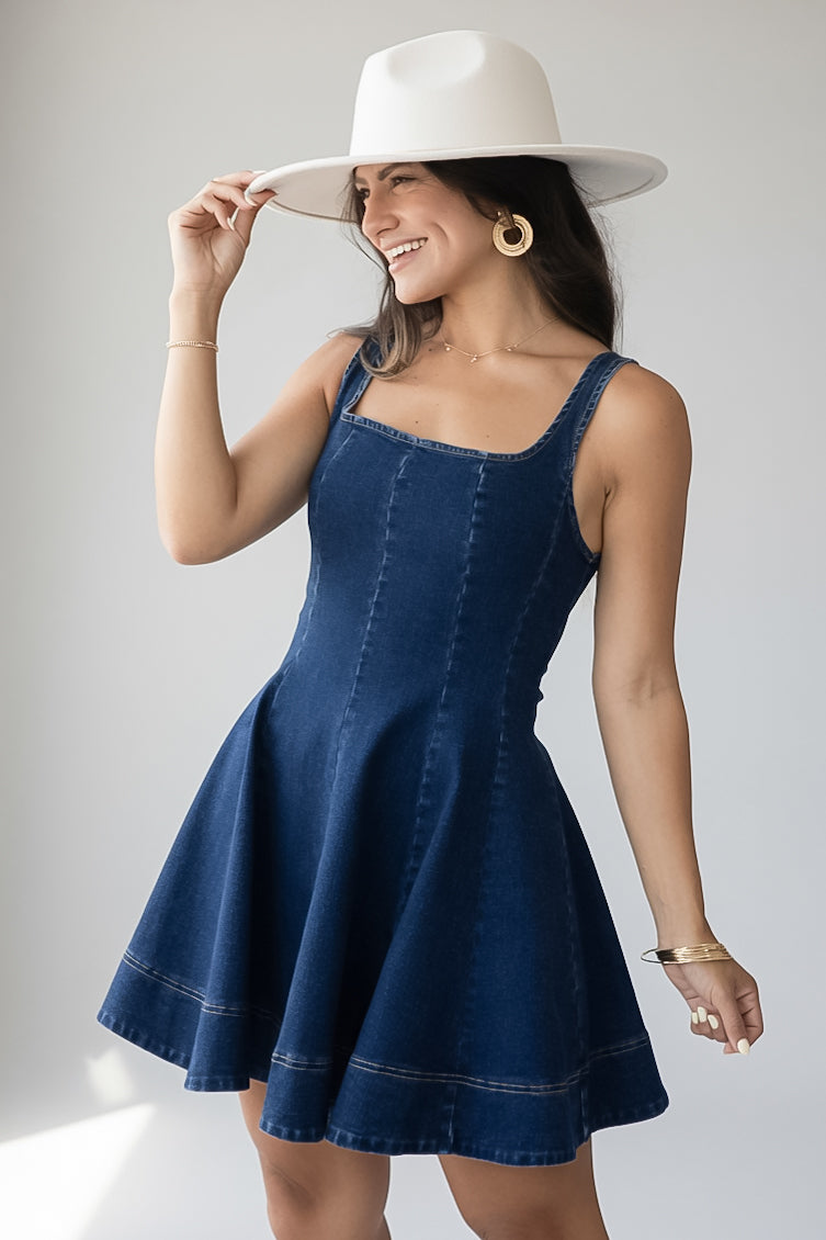 A woman wearing a dark blue denim pinafore dress with a flared skirt, styled with a wide-brimmed white hat, gold hoop earrings, and bracelets, posing against a light gray studio background.