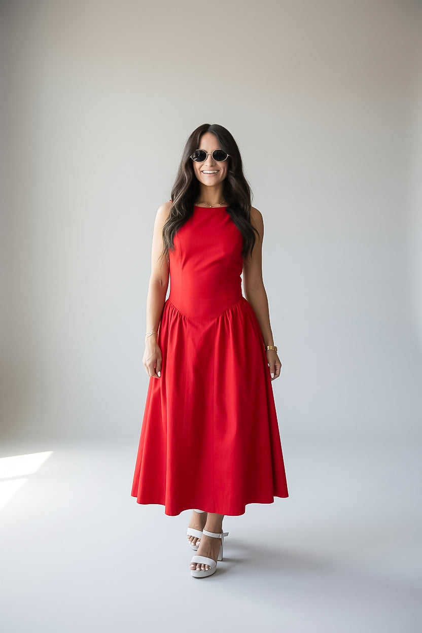 A woman modeling a sleeveless red midi dress with a fitted bodice and flared skirt, paired with white heeled sandals and sunglasses, standing against a plain light gray studio background.
