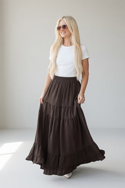 A blonde woman wearing a white short-sleeve t-shirt and a long, dark brown tiered maxi skirt with pockets, standing in a bright, minimalist studio.