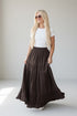 A blonde woman wearing a white short-sleeve t-shirt and a long, dark brown tiered maxi skirt with pockets, standing in a bright, minimalist studio.