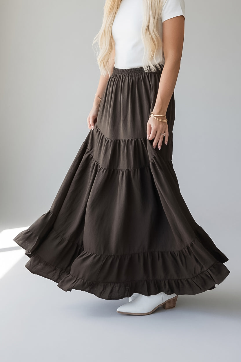 A woman wearing a dark brown, tiered maxi skirt with ruffled hems, paired with a white t-shirt and white heeled boots, shown from the waist down against a neutral gray background.