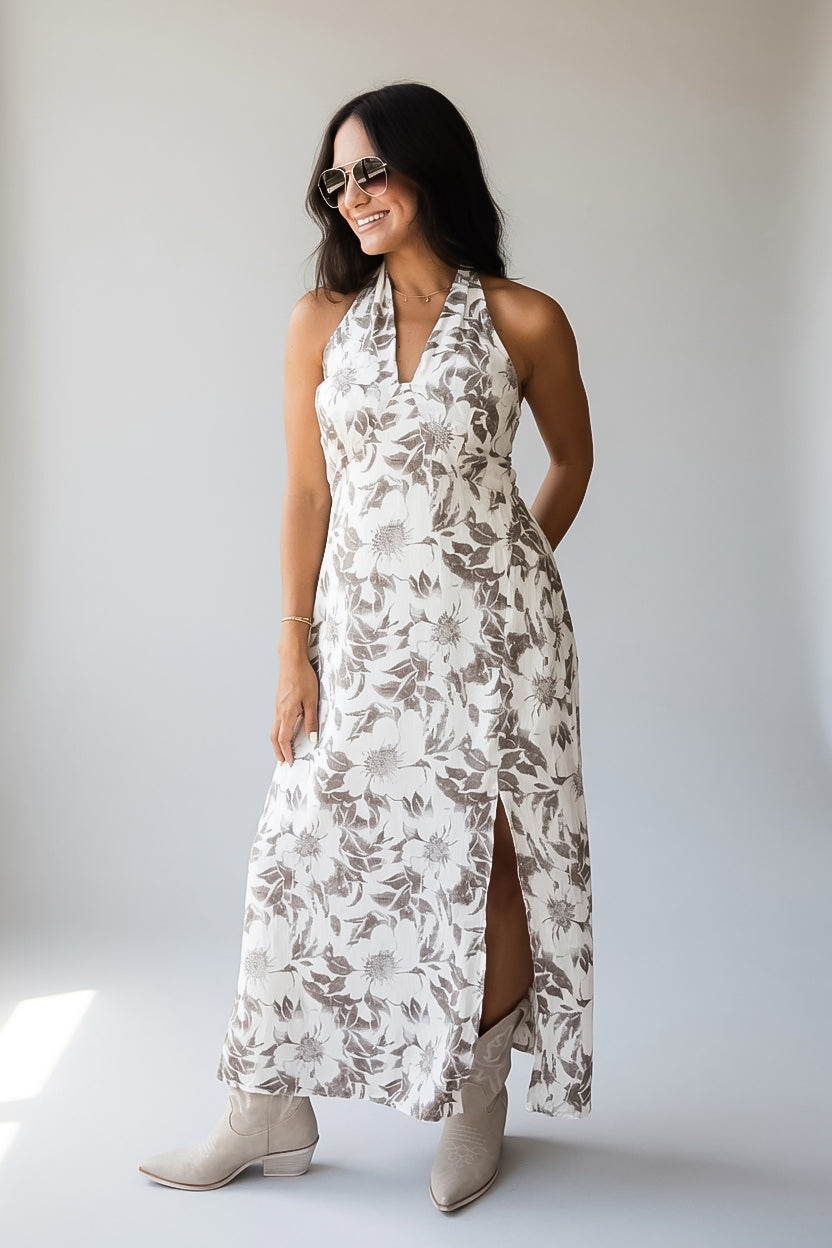 A woman modeling a sleeveless halter-neck maxi dress with a brown and beige floral pattern on a white background, paired with beige ankle boots, set against a plain light gray studio backdrop.