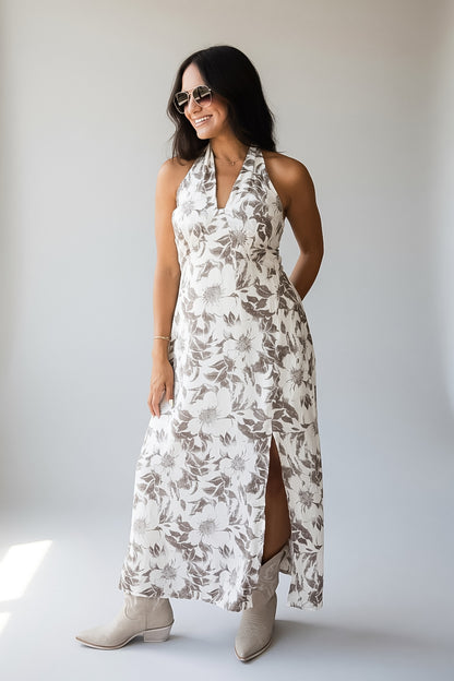 A woman modeling a sleeveless halter-neck maxi dress with a brown and beige floral pattern on a white background, paired with beige ankle boots, set against a plain light gray studio backdrop.