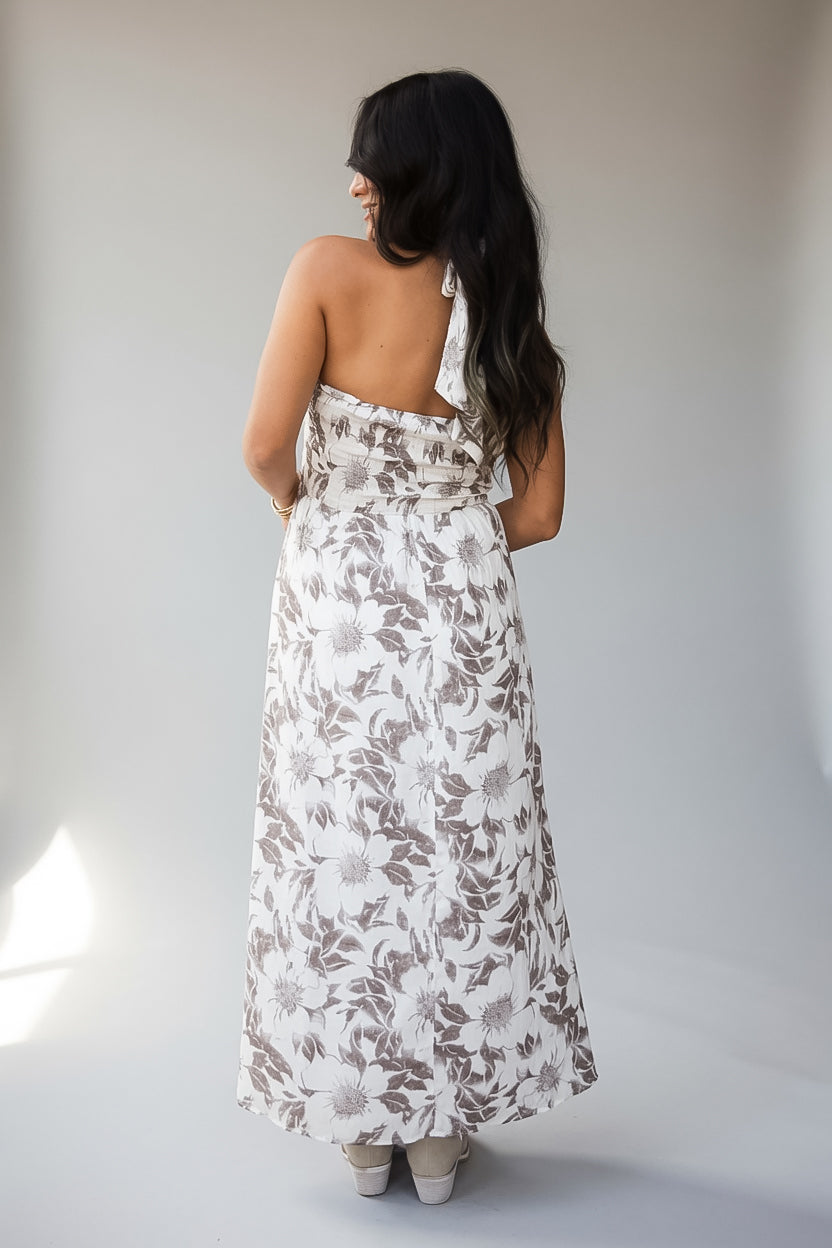 A woman viewed from behind wearing a sleeveless, backless halter-neck maxi dress with a brown and white floral pattern, standing against a neutral light gray studio background.
