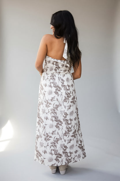 A woman viewed from behind wearing a sleeveless, backless halter-neck maxi dress with a brown and white floral pattern, standing against a neutral light gray studio background.