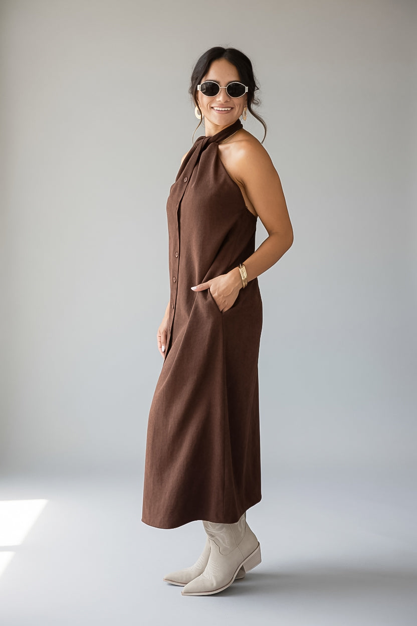 A woman modeling a sleeveless, high-neck brown midi dress with side pockets, paired with cream ankle boots and sunglasses, standing against a light gray studio background.