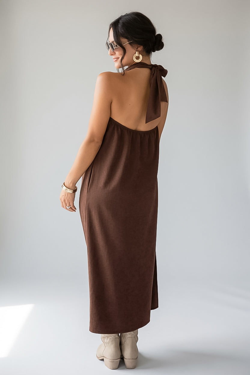 A woman wearing a brown halter-neck maxi dress with a tie-back detail, shown from the back against a light gray studio background.