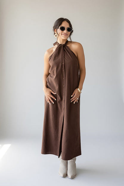 A woman modeling a sleeveless halter-neck midi dress in a solid dark brown color, featuring a button-down front and a front slit. She is wearing beige ankle boots, sunglasses, and a gold bracelet, standing against a plain light gray studio background.