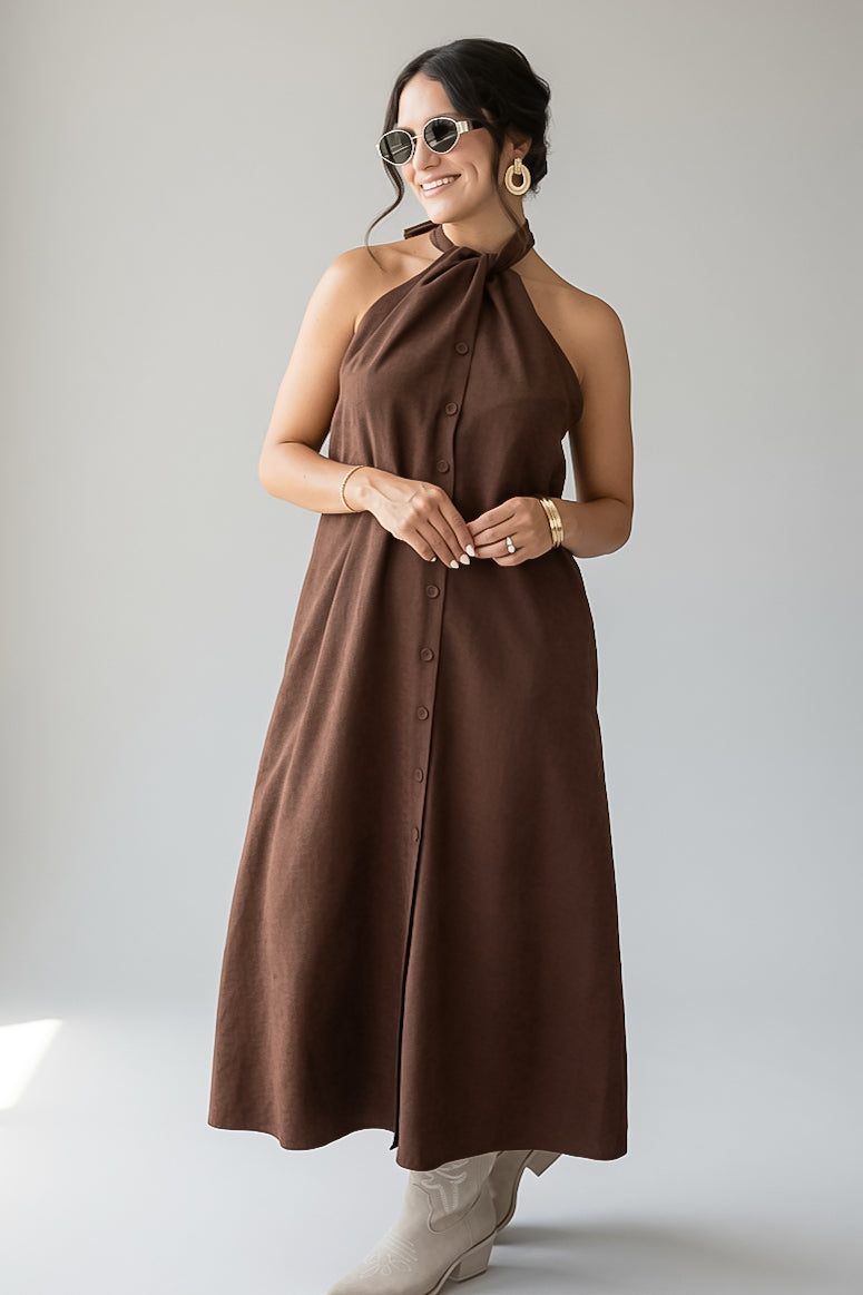 A woman modeling a brown halter-neck midi dress with a button-front closure, photographed against a plain light gray studio background.