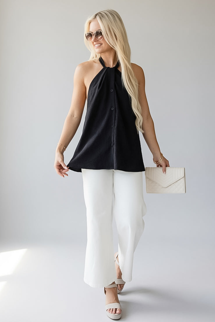 A blonde woman modeling a black sleeveless halter top with a button-front detail, paired with white wide-leg pants, white sandals, and a cream envelope clutch, set against a light gray studio background.