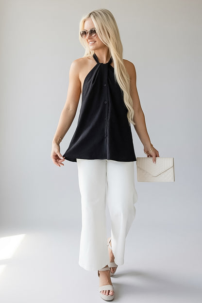 A blonde woman modeling a black sleeveless halter top with a button-front detail, paired with white wide-leg pants, white sandals, and a cream envelope clutch, set against a light gray studio background.