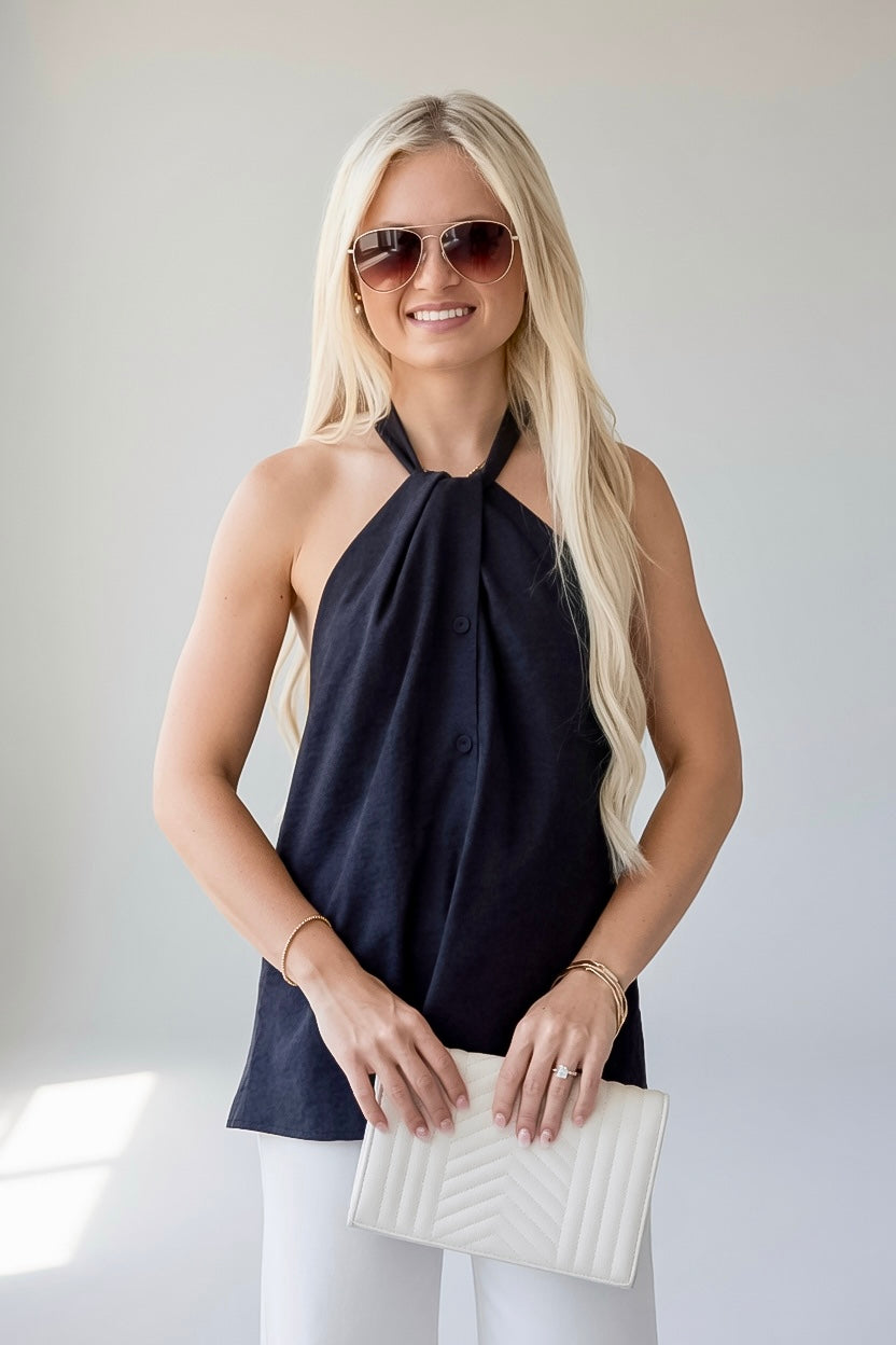 A blonde woman wearing a navy blue halter top, white pants, and sunglasses, holding a white quilted clutch bag.