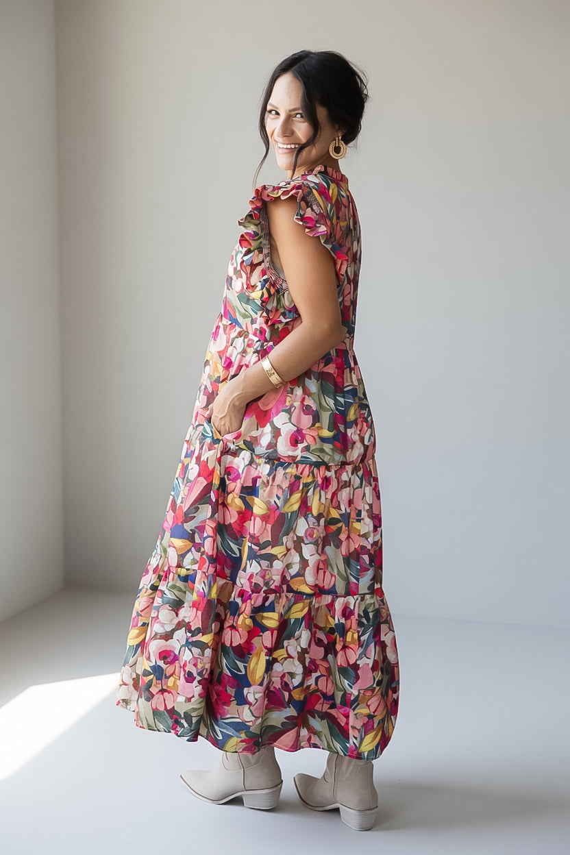 A woman wearing a colorful floral tiered maxi dress with ruffled sleeves, paired with light beige ankle boots, standing in a minimalist studio setting.