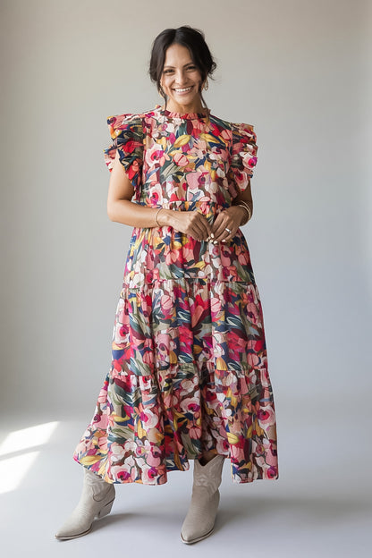 A woman modeling a colorful floral midi dress with ruffled short sleeves and a tiered skirt, paired with beige ankle boots, against a neutral studio background.
