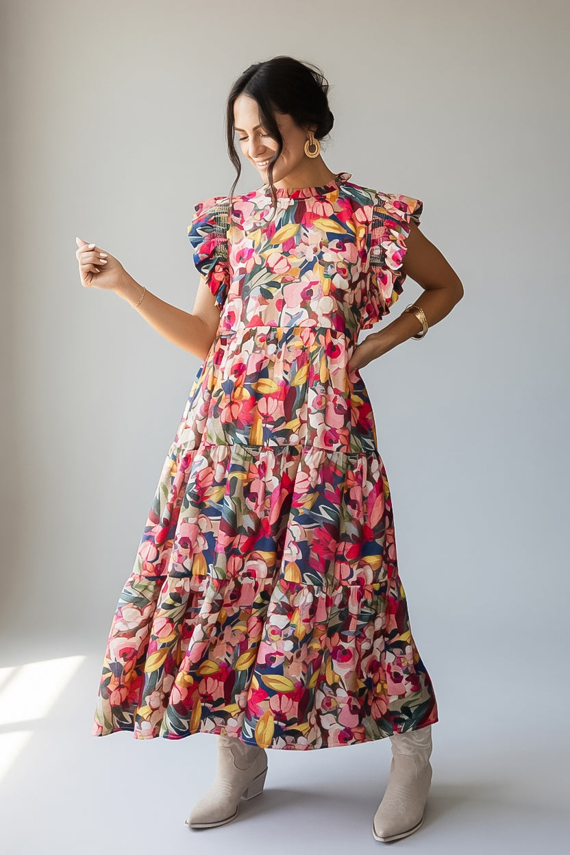 A woman modeling a tiered, short-sleeved maxi dress with a vibrant multicolor floral print, paired with beige ankle boots, set against a plain light gray studio background.