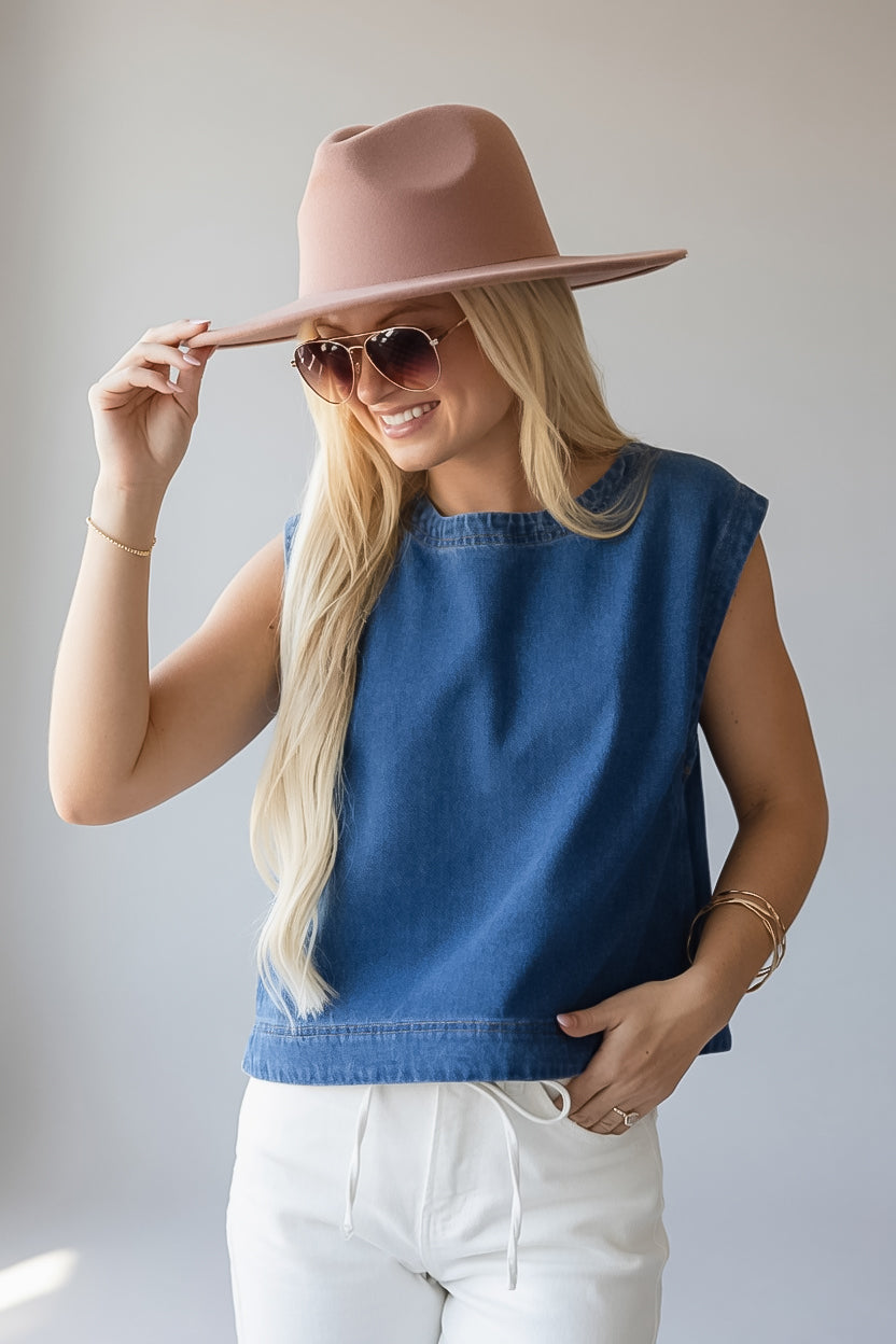 A woman wearing a blue sleeveless top, white pants, a dusty pink wide-brim hat, and sunglasses, posing against a light gray background.