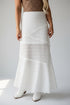 A woman wearing a white textured lace maxi skirt with a ruffled hem, paired with white pointed-toe boots against a light gray studio background.