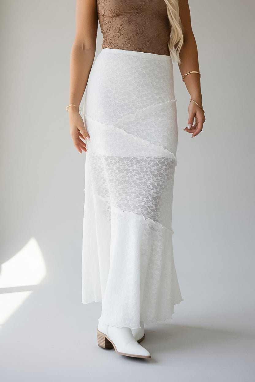 A woman modeling a white textured wrap-style maxi skirt with layered ruffle details and sheer patterned sections, paired with white pointed-toe boots against a light gray studio background.