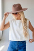A woman wearing a white sleeveless cropped top, blue jeans, a brown wide-brim hat, and sunglasses, posing in a bright indoor setting.