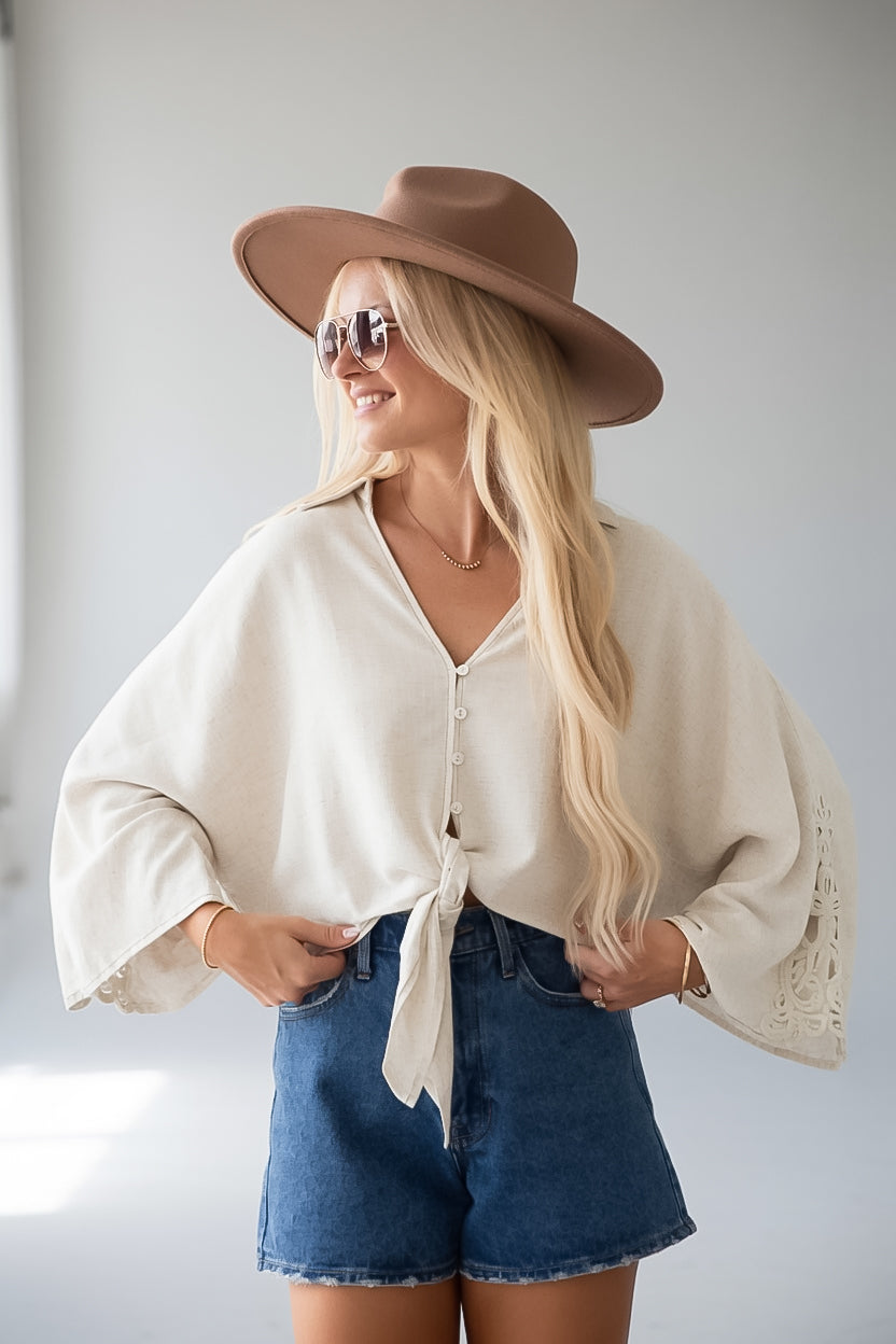 A woman wearing a cream-colored bohemian-style tie-front blouse with dramatic bell sleeves, blue denim shorts, a brown wide-brim hat, and sunglasses, posing against a light gray background.