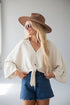 A woman wearing a cream-colored bohemian-style tie-front blouse with dramatic bell sleeves, blue denim shorts, a brown wide-brim hat, and sunglasses, posing against a light gray background.