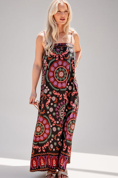 A woman modeling a sleeveless maxi dress with thin straps and a vibrant bohemian mandala print on a black background, set against a neutral gray studio backdrop.