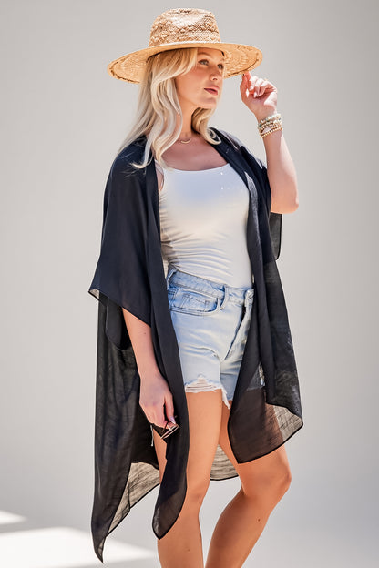 A woman modeling a sheer black open-front kimono-style cover-up over a white tank top and light denim shorts, wearing a straw hat against a plain light gray studio background.