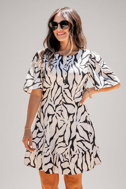 A woman modeling a short-sleeve ruffled mini dress with a black and white abstract floral pattern, styled with black sunglasses against a solid light gray background.
