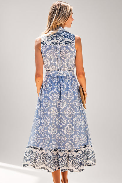 A woman modeling a sleeveless midi dress from the back, featuring a light blue and white damask pattern with black lace trim at the high neckline, waist, and ruffled hem, set against a plain light gray studio background.