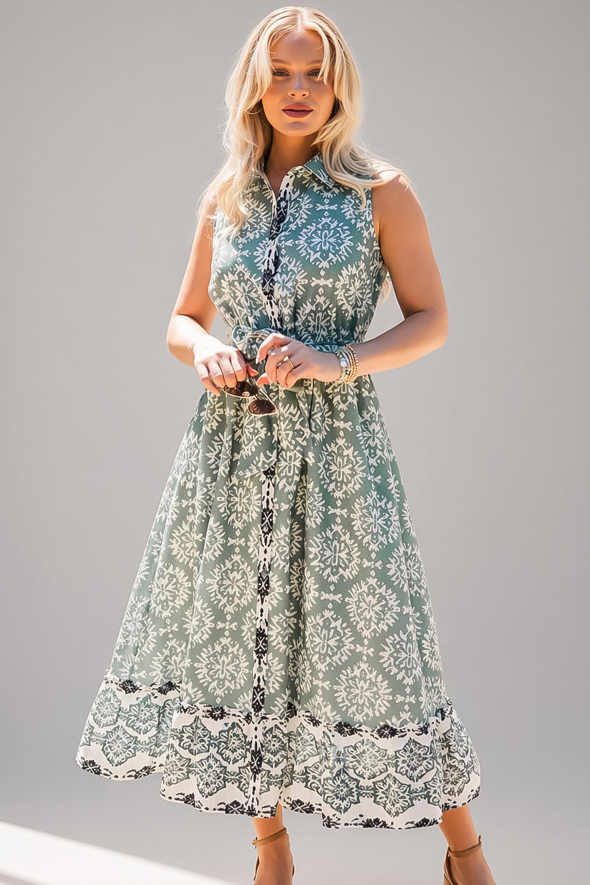 A woman modeling a sleeveless midi dress with a green and white floral damask pattern, featuring a button-up front and a ruffled lace hem, set against a plain gray studio background.