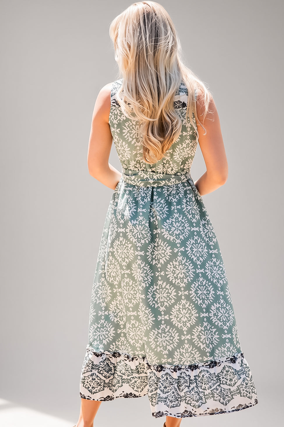 A woman viewed from behind wearing a sleeveless midi dress with a white floral damask pattern on a sage green background and a black lace hem, set against a plain gray studio background.