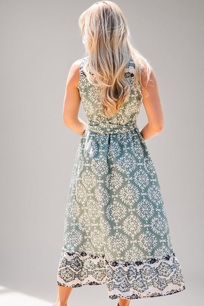 A woman viewed from behind wearing a sleeveless midi dress with a white floral damask pattern on a sage green background and a black lace hem, set against a plain gray studio background.