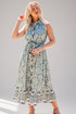 A woman modeling a sleeveless midi dress with a sage green and white floral pattern, featuring a button-up front, tie waist, and ruffled hem, set against a plain light gray studio background.